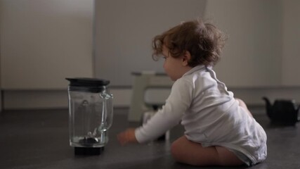 Curious toddler reaching toward upright blender jar on kitchen floor while crawling with focused intent during gentle moment of exploration as family moves through background