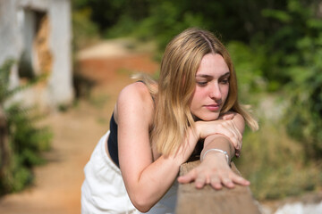 Portrait of young, beautiful, blonde woman in black T-shirt and white pants, looking distracted, leaning on a wooden railing in an outdoor park. Concept beauty, youth, millennial.