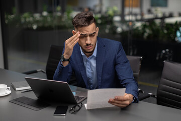In a contemporary office setting, a sad middle eastern manager sits at his worktable, reading a marketing report. He is dressed in a suit, looking worried and touching his forehead, indicating stress.