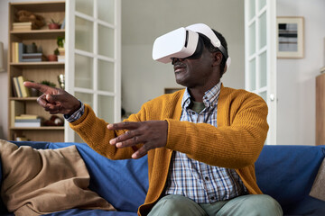 Mature Black man sitting on sofa using virtual reality headset, extending arms and interacting with digital environment, appearing engaged with immersive technology experience