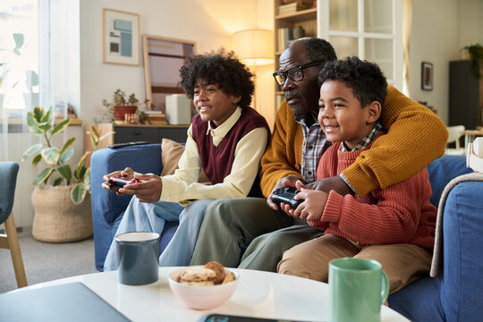Black senior man sitting on sofa playing video game with two Black boys, smiling and holding controllers, spending quality time together in modern living room at home