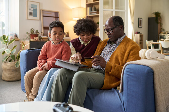 Black senior man sitting on sofa with two Black children, holding credit card and showing tablet, children attentively watching screen, family engaging in online shopping together