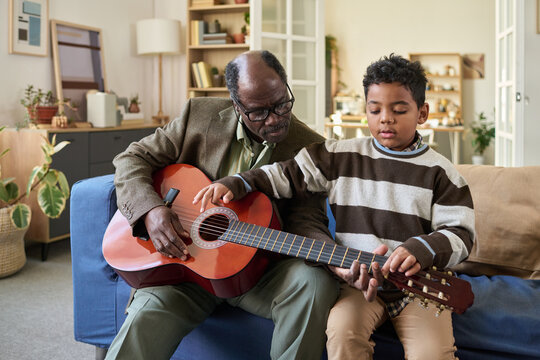 Senior Black man teaching child boy guitar, sitting together on sofa, focusing on hand placement and technique, intergenerational music lesson in home living room