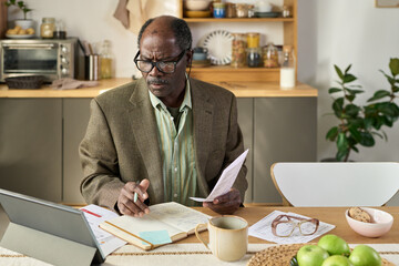 Senior Black man wearing glasses sitting at kitchen table analyzing financial documents and writing notes in notebook while using digital tablet