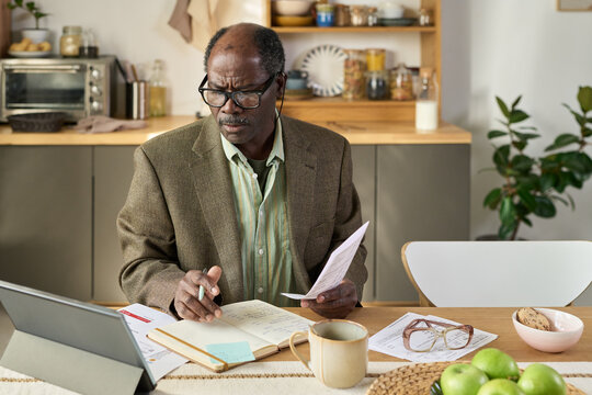 Senior Black man wearing glasses sitting at kitchen table analyzing financial documents and writing notes in notebook while using digital tablet