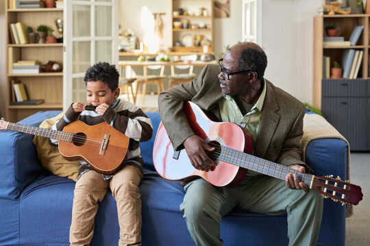 Senior man sitting on sofa playing acoustic guitar while looking at Black child holding ukulele, both engaging in music activity together in cozy living room setting - Powered by Adobe