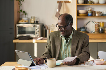 Senior Black man sitting at table using digital tablet and holding paperwork, smiling while managing finances or reviewing documents in kitchen setting, coffee mug nearby