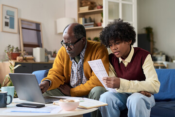 Mature Black man sitting with teenage Black boy using laptop and reviewing financial documents together, both focusing on paperwork in modern living room setting