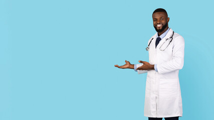 Medical Ad. Smiling black male doctor pointing aside at copy space, young african american physician wearing uniform standing isolated over blue background, panorama with free place for design