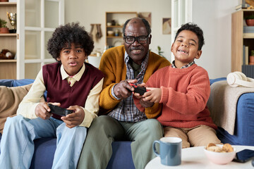 Senior Black man sitting between two Black boys playing video games together on couch, all holding controllers and smiling, multigenerational family bonding in living room setting