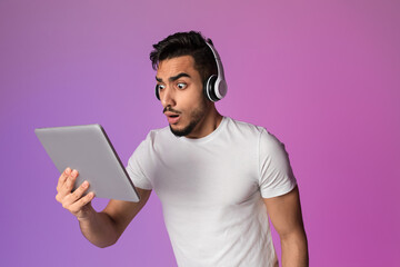 A shocked Arab man in headphones stares at his tablet in disbelief. Bright neon lights surround him as he realizes he has won a significant online lottery or casino bet.