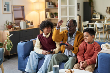 Black senior man sitting on sofa playing video game with two Black boys, one teenager and one child, all holding controllers and focusing on television screen in modern living room