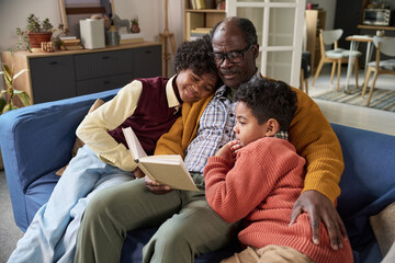 Obraz premium Senior Black man sitting on sofa reading book to two Black children, both kids leaning against him and listening attentively, cozy family moment in home living room setting