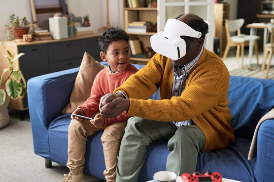 Senior Black man wearing virtual reality headset interacting with digital environment while sitting on sofa next to Black boy watching attentively
