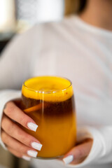 A close-up shot of a person holding a glass with a vibrant, two-toned layered beverage, possibly orange juice and coffee, featuring a bright yellow top layer and dark bottom layer.