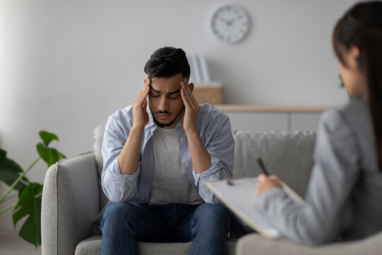 A stressed middle eastern male patient sits on a couch in a psychologist's office. He holds his head in his hands, expressing feelings of anxiety and depression, while a therapist takes notes.