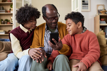Senior Black man sitting between two Black boys showing smartphone screen, boys attentively watching device, family members interacting closely on couch in home setting