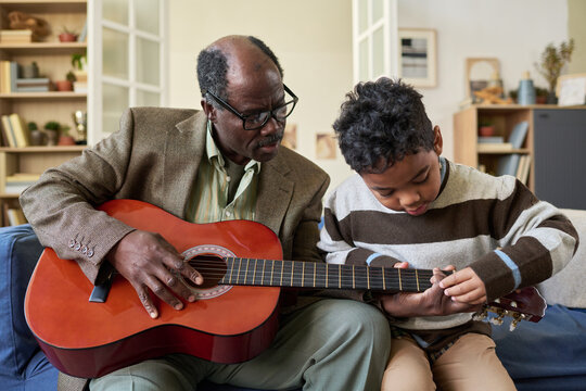 Black senior man teaching Black child playing acoustic guitar together on sofa, both focusing on instrument, intergenerational bonding, home setting, music education concept