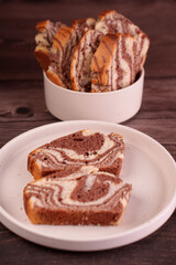 close-up view of delicious sliced marble cake on a white plate and full bowl against dark wooden background, highlighting swirled chocolate and vanilla patterns, comfort, confection, toppings