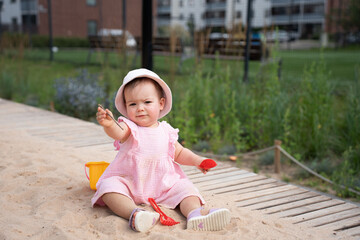 adorable toddler in pink dress and sun hat playing with toy shovel and bucket in sandbox on warm...