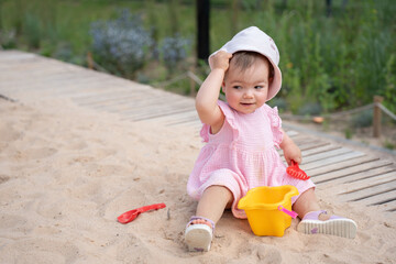 adorable toddler in pink dress playing joyfully in a sandy playground, holding a red toy rake and yellow bucket, set against lush green background and wooden pathway, park, relaxed, cheerful