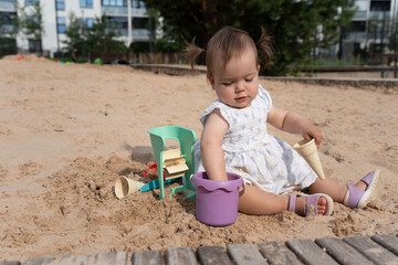 young child in white dress plays with purple bucket and toy in outdoor sandbox, surrounded by greenery and urban buildings, on a sunny day, blue, innocent, explore, adventure, carefree, childhood