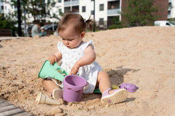 young child in a white dress plays in a sunlit sandbox with colorful plastic toys under clear skies, surrounded by greenery and urban buildings in the background, happiness, care, growth, adventure
