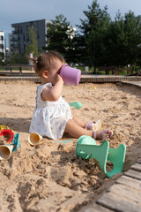 toddler in a white dress plays with colorful toys in a sandbox, enjoying a sunny day in an urban park with surrounding greenery and modern buildings, development, interaction, joy, happiness