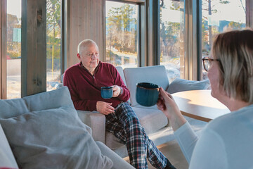Calm morning exchange as a senior couple sits with warm mugs in soft Nordic daylight, sharing a slow conversation with steady presence. Gentle tones, quiet connection , copy space.