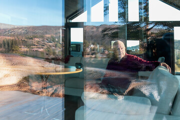Reflective stillness as a senior man sits inside a Nordic cabin while the landscape blends with his silhouette in the window. Layered textures, quiet awareness and calm moment of pause,copy space .