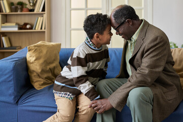 Black senior man sitting on sofa touching foreheads with boy smiling warmly, both holding hands and sharing affectionate moment in cozy living room setting