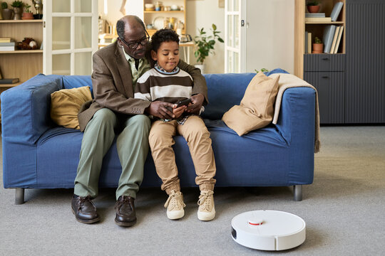 Senior Black man sitting on sofa with Black boy using digital tablet together, both looking at screen while robotic vacuum cleaner moving on floor in modern living room