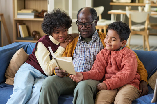 Black senior man sitting on sofa reading book to two Black children, one girl and one boy, both leaning in and listening attentively, family spending time together indoors