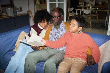 Black mature man sitting on sofa reading book with two Black boys, one child and one teenager, both listening attentively and engaging with story in cozy home living room setting