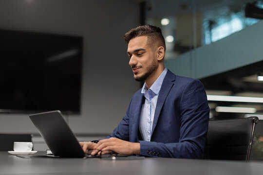 A young middle eastern businessman sits at a sleek worktable in a modern office, focused on typing on his laptop. He is engaged in sending emails and conducting marketing research.