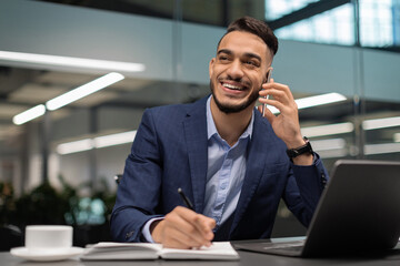A young arab businessman in a blue suit sits at his worktable in a modern office. He is talking on his mobile phone, taking notes, and smiling as he engages with his work and surroundings.