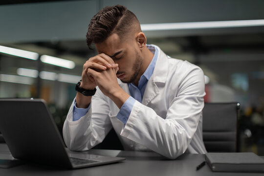 Middle eastern young man in white workwear sits at a desk, looking exhausted while facing a laptop. He seems to be feeling the pressure of work and in need of a break or vacation.