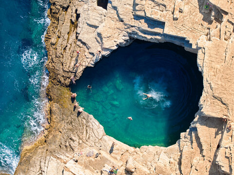 Top-down drone of Giola rock pool on Thassos with swimmers and a fresh splash in emerald water. Ideal for: Greece travel, adventure tourism, summer campaigns, social ads. Colours: teal, blue, beige.