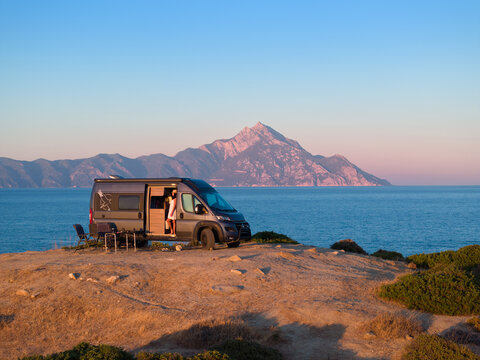 Woman enjoys the Aegean view beside a camper van with Mount Athos in the background. Ideal for: vanlife travel, outdoor brands, road-trip blogs, Greece tourism. Colours: blue, beige, silver, gold.