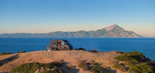 Woman enjoys the Aegean view beside a camper van with Mount Athos in the background. Ideal for: vanlife travel, outdoor brands, road-trip blogs, Greece tourism. Colours: blue, beige, silver, gold.