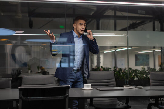 A young middle eastern businessman walks past a modern office with glass walls, gesturing wildly during a phone call with a business partner. He appears frustrated as he talks and looks away.
