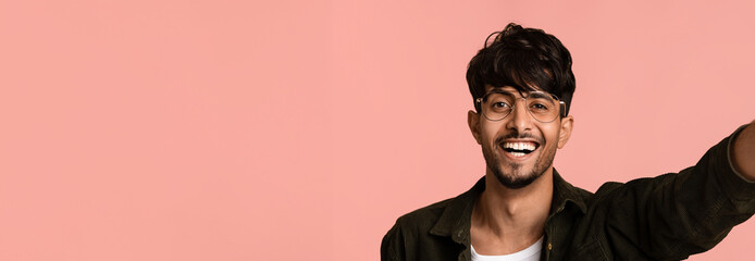 A young man with glasses and a stylish outfit is smiling broadly and waving. He stands against a soft pink background, radiating joy and positivity in a casual environment.