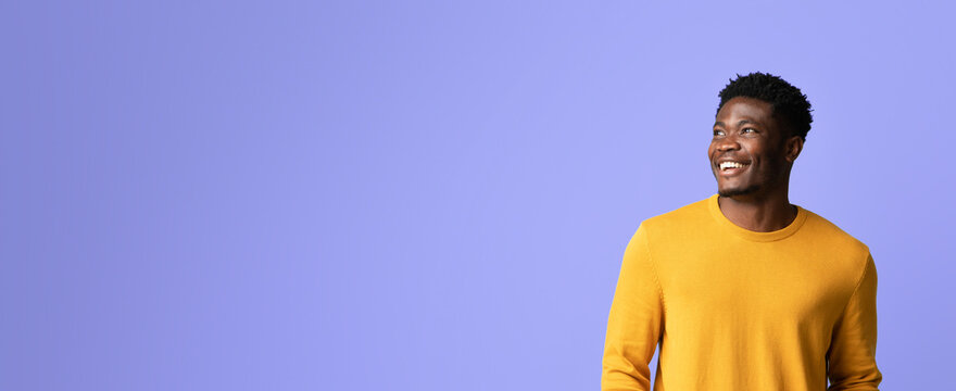 A man with a joyful expression stands against a purple wall. He is wearing a bright yellow sweater and appears happy, enjoying a light-hearted moment. The setting is bright and uplifting.