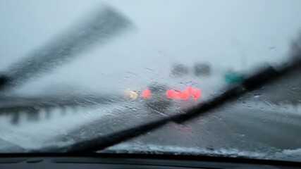 driving in bad winter weather with sleet and snow on windshield. wiper blade clears glass, revealing blurry car taillights ahead. poor visibility on dangerous road.