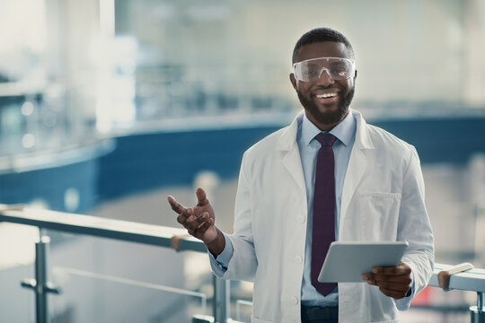 Handsome young african american man scientist in workwear and protective glasses standing with digital tablet at laboratory, smiling and gesturing, copy space. Modern technologies in science