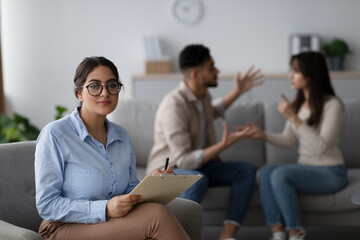 In a counseling office, a serious family counselor looks at the camera while a young couple argues...