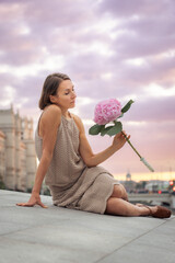 Woman in a beige knitted dress sits gracefully on a stone surface, holding a pink hydrangea against a pastel sunset sky