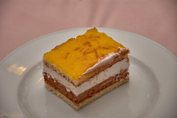 Slice of cake with layers of cream, chocolate, and yellow frosting in the foreground on a white plate in a food scene.