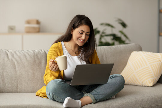 A happy arab female freelancer sits on her sofa, sipping coffee from a cup. She is engaged with her laptop, working online in a bright and inviting living room space. - Powered by Adobe