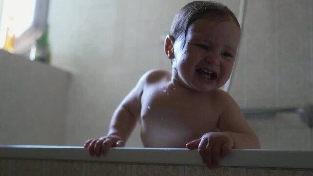 Baby crying while standing in bathtub with water droplets on skin, emotional moment showing discomfort, vulnerability, need for reassurance, and raw expression during bath time routine